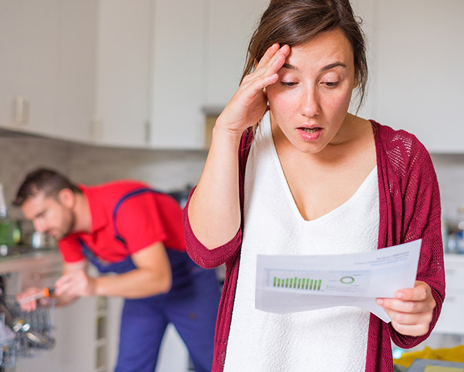 A repair man fixes a dishwasher behind a woman who is shocked her claim is denied.