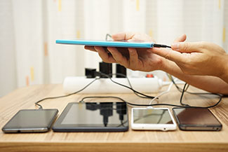Image shows a person plugging a phone into charger, where four other phones are also charging via a surge protector.