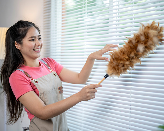 A smiling woman cleans her white window blinds with a feather duster.