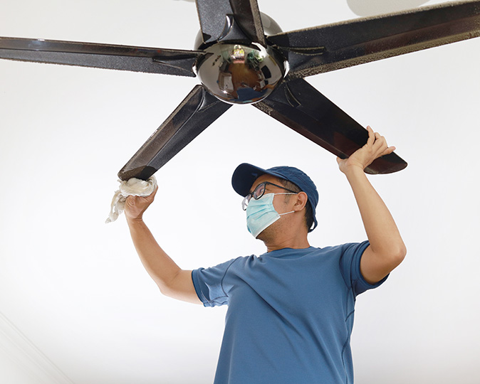A man in blue shirt and face mask cleans a ceiling fan at home.