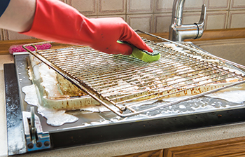 A person wearing orange rubber gloves scrubs an oven rack next to the sink with a green sponge and soapy water