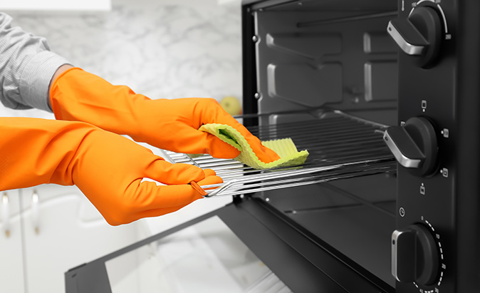 A closeup of a homeowner’s hands in orange rubber gloves uses a yellow sponge to clean the racks in the oven.