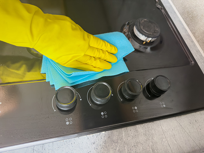 Person wearing yellow rubber glove uses cloth to clean around gas stove burners.