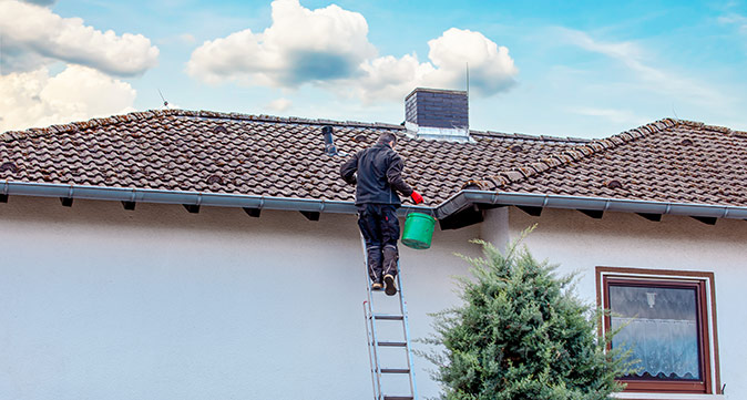 A man climbing a ladder leaning against a house and carrying a green bucket to clear debris from plumbing vent pipe.
