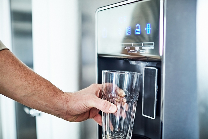 The arm of an out-of-frame man holds an empty glass up to the water dispenser of a modern refrigerator.