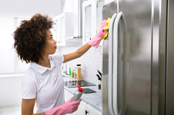 A homeowner wearing pink rubber dish gloves polishes her stainless steel refrigerator with a sponge in a brightly lit kitchen.