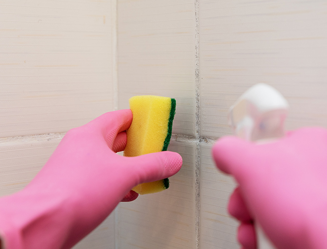 A homeowner wearing rubber gloves and holding a spray bottle sprays cleaner on shower mold.