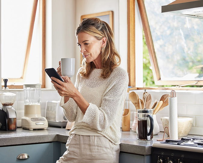 A woman holding a mug in her kitchen prepares to make a call to a repair person.