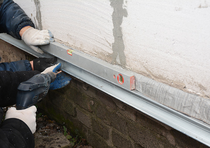 Two contractors using a level to install metal sheets for waterproofing the foundation on the outside of a house.