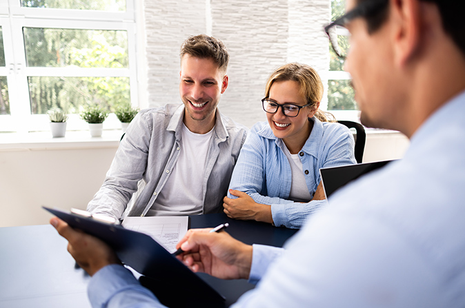 A young couple sitting at a table with a financial consultant pointing at a clipboard.