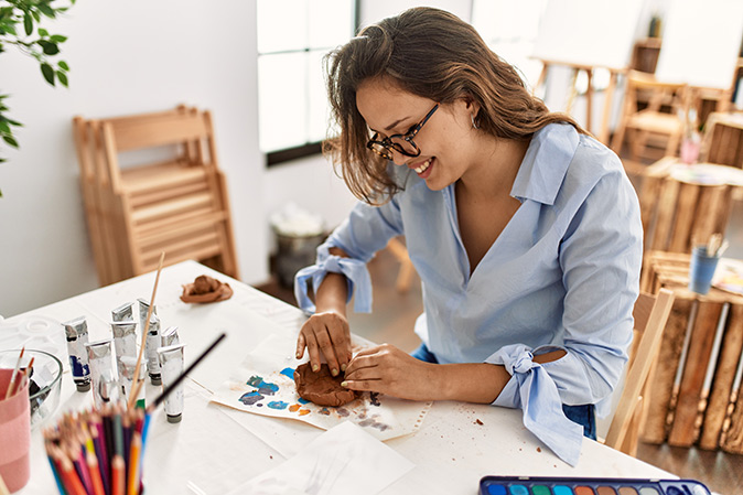 A woman in a blue shirt handling clay at a desk in a craft room.