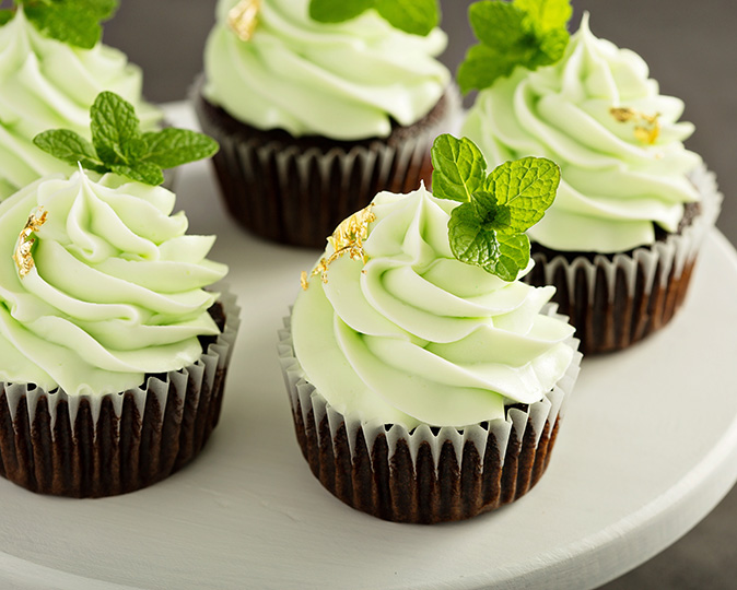 A white circular tray displays festive mint chocolate cupcakes with gold leaf and mint garnish.