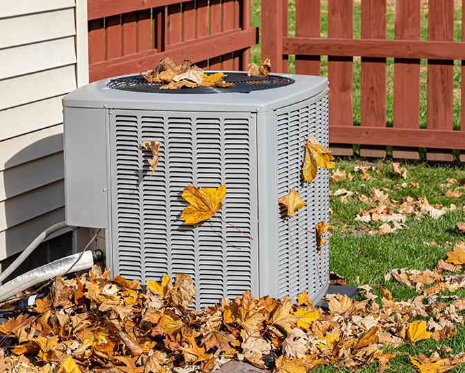 An AC unit sits in a yard covered in and surrounded by autumn leaves.