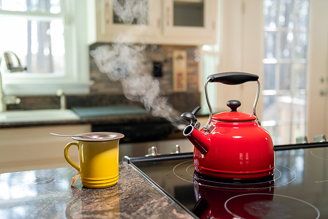 An electric stove heating up a red tea kettle, with a yellow cup sitting to its left.