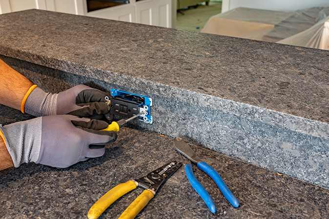A homeowner replaces an old outlet on a granite kitchen island with a black GFCI outlet