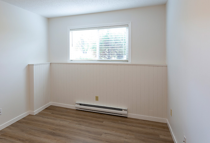 A small, empty white room with an overhead light and window that has a small baseboard heater installed on one wall below the window.