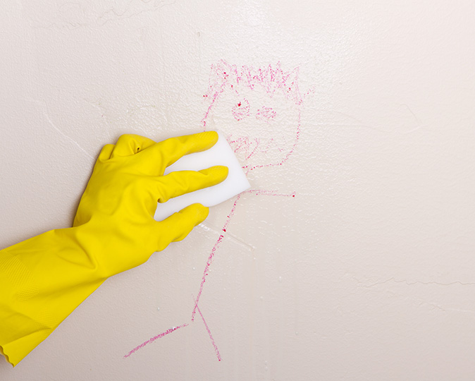 A gloved hand wipes a wall marked with a crayon drawing with a magic eraser sponge.