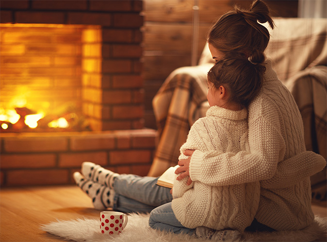 A mother and child cozy up to a fireplace with a book and tea cup in their living room.