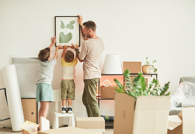 Surrounded by moving boxes, a father and his young sons hang a picture in their new home.