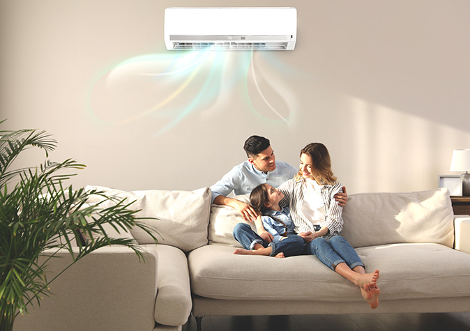A family of three sitting on a beige couch under a wall-mounted air conditioning unit.