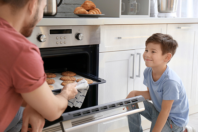 A young boy watches his father pull freshly baked cookies from their electric oven. 