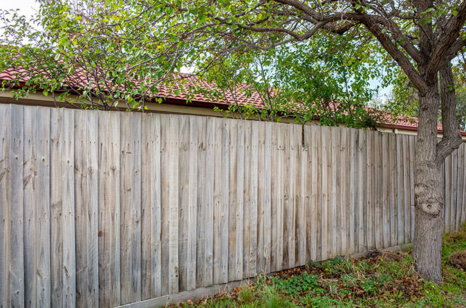 A weathered wood fence separates one home’s backyard from another where a red tile roof is visible above the fence line.