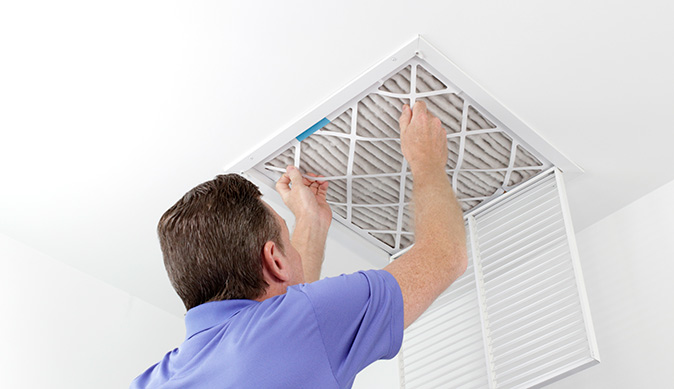 A homeowner reaches up to remove a used air filter from an open central heating vent.