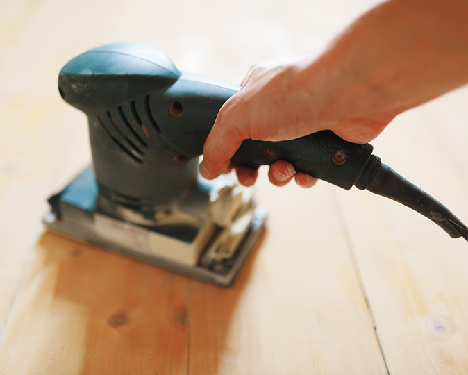 A homeowner sands a hardwood floor with a handheld power sander.