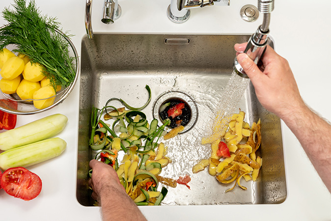 A homeowner peels a zucchini with a vegetable peeler as the food scraps collect in the sink.