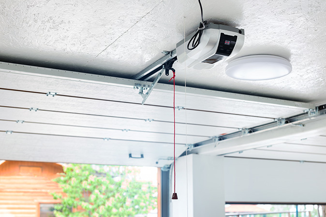 An automatic garage door opener mounted on the ceiling with a red emergency cord and a rolling garage door opening.
