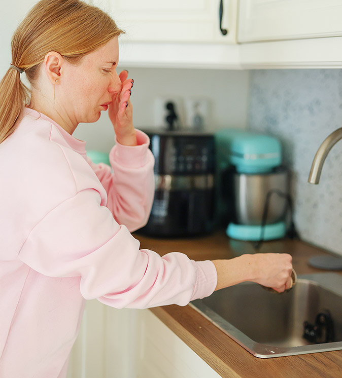 A woman trying not to smell something unpleasant coming from the kitchen sink.