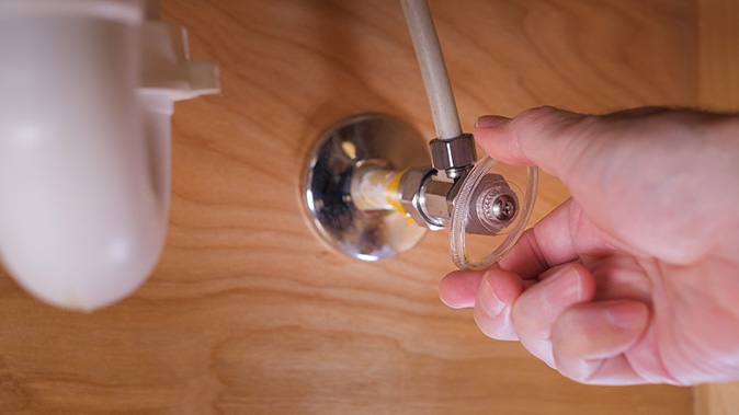 A person’s hand turning off the water shut-off valve under a sink.