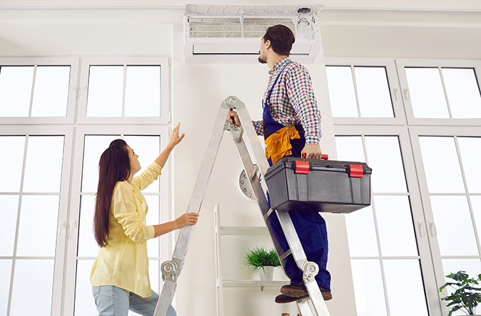 A technician climbs a latter to repair a faulty air conditioner.