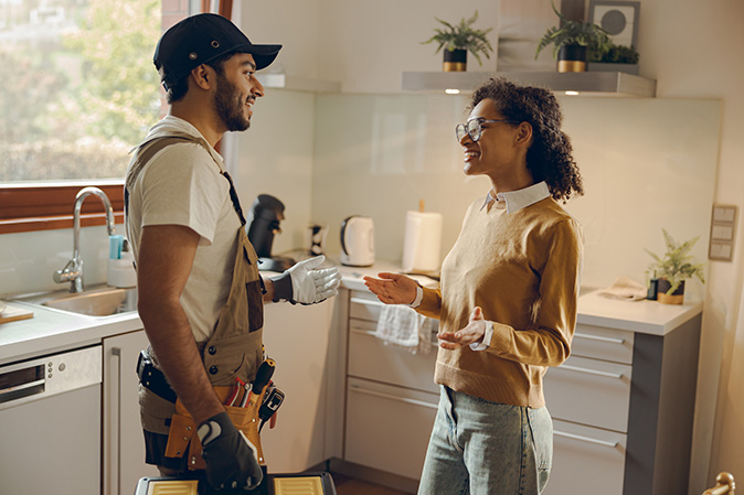 A handyman standing in a kitchen and talking with a homeowner about the issue that needs repairs.