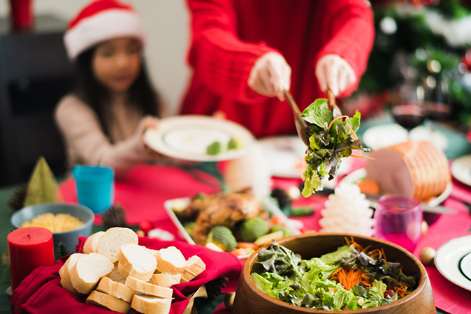 A mother dressed in Christmas red helps scoop salad greens for her daughter, who is wearing a Santa hat.