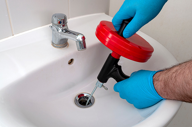 A homeowner uses a drain snake to unclog the sink while wearing rubber gloves.