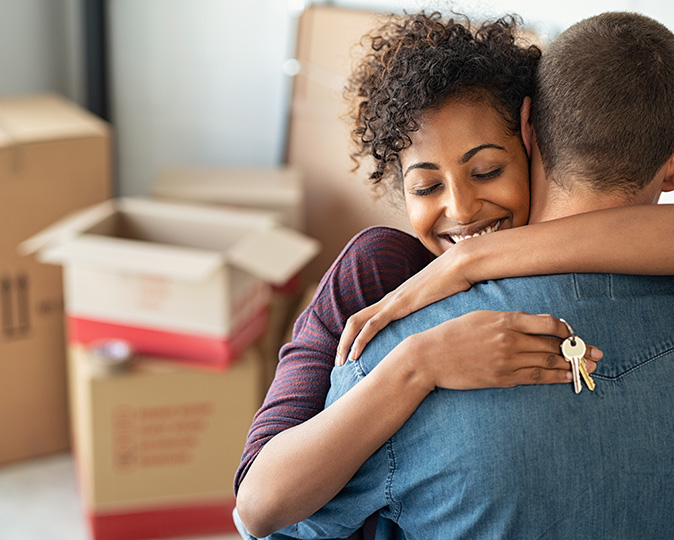 A young couple hugs, holding the keys to their new home while packing up their old home.