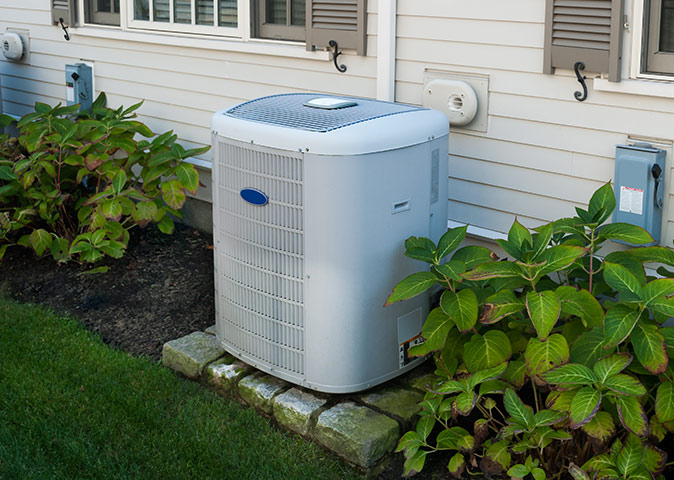 A central air conditioner against the side of a house by plants.