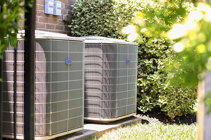 Two outdoor HVAC units on a concrete block in the summer, surrounded by greenery