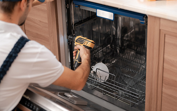 A person using a screwdriver to secure a dishwasher to the neighboring cabinets.