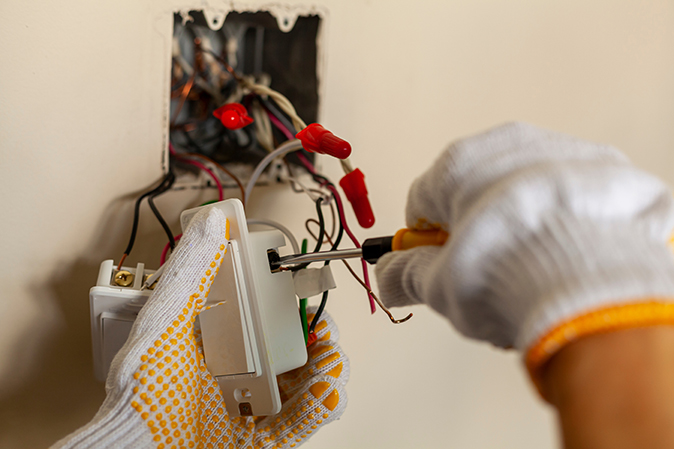 A close-up of a person’s hands using a screwdriver to wire a connection to a light switch.