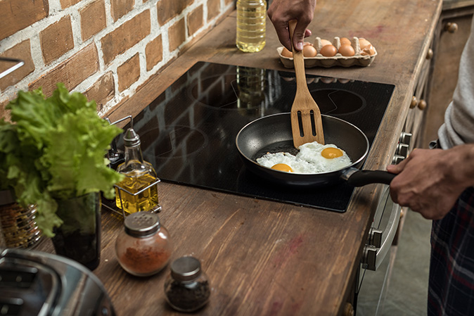 A man out of frame frying two eggs in a pan on an induction stovetop surrounded by oils and seasonings in the background.
