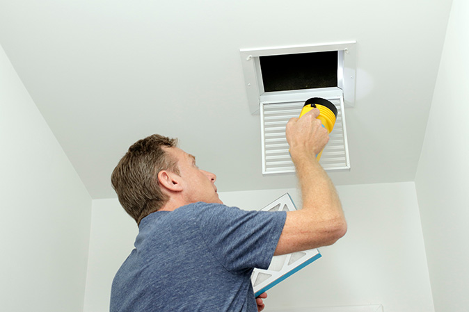 A man with a yellow flashlight inspects the inside of an air vent.