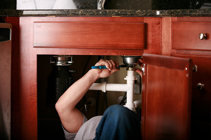 A homeowner works underneath a kitchen sink to detach pipes with straight jaw pliers and replace the garbage disposal.