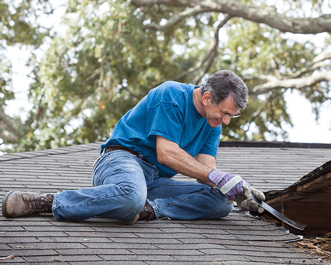 A man wearing gloves attempting repairs to his worn, leaking roof with a prybar.