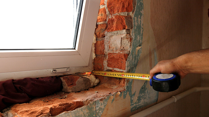 A person’s hand using a measuring tape to measure the depth of a window against a brick wall.