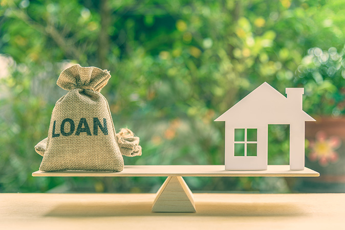 A loan bag balances perfectly with a house model on a basic balance scale.
