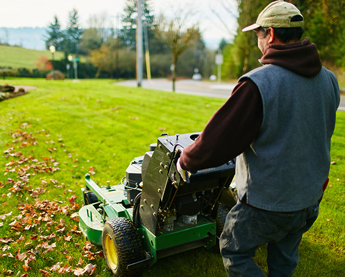 A homeowner with a large lawnmower mows a large front yard scattered with leaves.
