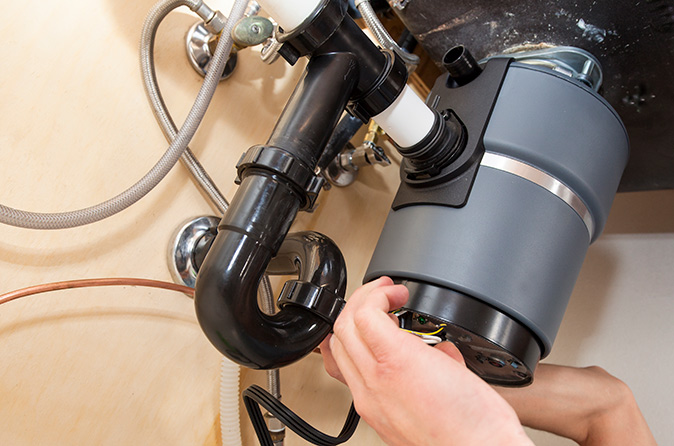 Under a kitchen sink, a person’s hand holds an orange wrench and tightens their sink drain.