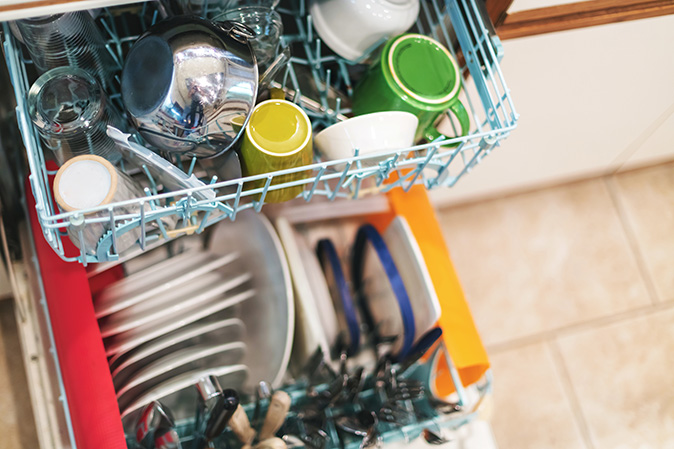 An open, under-the-counter dishwasher set into cabinet is overloaded with dishes, bowls, and utensils.